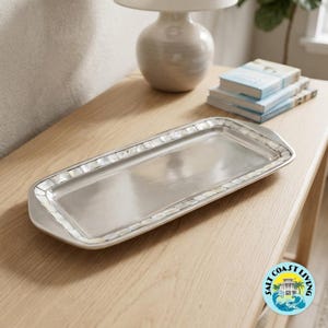 May include: A rectangular silver-toned metal serving tray featuring a smooth, polished center surface and a decorative border inlaid with small, iridescent white shell segments. The tray has integrated handles on both short ends.