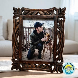 May include: A dark brown wooden picture frame with intricate carved leaf patterns along the border. The frame holds a photograph of a person in a dark jacket and baseball cap kneeling on a deck while interacting with a brown and white dog.