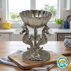 May include: A metallic silver decorative bowl featuring a scalloped shell-shaped top supported by two seahorse figures standing on a circular base. The surface has a reflective finish, and the structure is designed as a pedestal centerpiece.