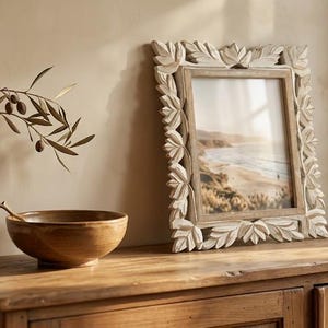 May include: A square picture frame with a white carved leaf border containing a coastal landscape print, positioned next to a brown ceramic bowl holding a single olive branch with small dark olives and green leaves.
