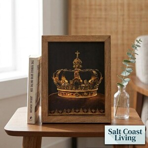 May include: A framed print featuring a detailed illustration of a golden crown with intricate metalwork and embedded jewels, set against a dark background. The crown rests on a dark cushion, and the artwork is encased in a light brown wooden frame.