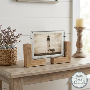 May include: A rectangular glass picture frame held by two L-shaped light brown wooden stands. The frame displays a sepia-toned photograph of a lighthouse. The frame is positioned horizontally, featuring a thin silver metal border around the glass.