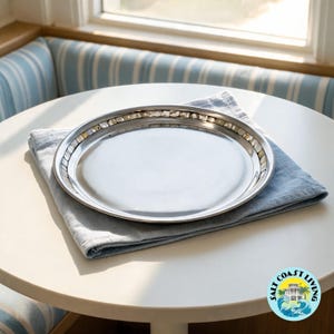 May include: A round, polished silver-toned metal serving tray featuring a decorative rim inlaid with small, square, iridescent mother-of-pearl tiles. The tray has a smooth, reflective surface and a raised edge, presenting a clean and metallic aesthetic.