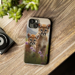 Könnte beinhalten: Eine Handyhülle mit einem Foto von zwei Pumas, einem erwachsenen Tier und einem Jungtier, die in einem Feld mit lila Blumen stehen. Der erwachsene Puma schaut in die Kamera, während das Jungtier zur Seite schaut. Der Hintergrund ist ein verschwommenes Bild einer Bergkette.