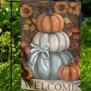 May include: A garden flag with a design of stacked pumpkins in orange, white, and grey, with a white bow. Sunflowers and autumn leaves surround the pumpkins. The word "WELCOME" is printed at the bottom of the flag.
