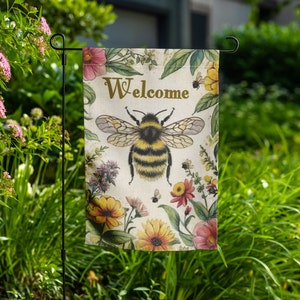 May include: A welcome garden flag featuring a bee and floral design. The flag is white with a yellow bee and colourful flowers. The text "Welcome" is written in gold lettering.