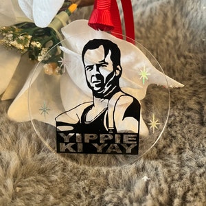May include: A clear round Christmas bauble featuring a black and white image of a man with the text "YIPPIE KI YAY" printed below. The bauble has a red ribbon attached to the top.