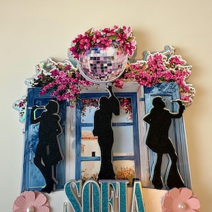 May include: A decorative cake topper featuring three black silhouettes of figures in flared outfits standing before a blue window frame. Above, a disco ball is surrounded by pink flowers. The name SOFIA is in teal glittery letters at the base.