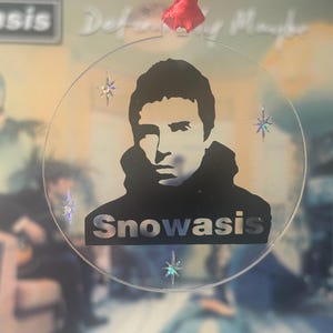Könnte beinhalten: Ein klarer, kreisförmiger Weihnachtsschmuck mit einer schwarzen Silhouette eines Männergesichts und dem Wort "Snowasis" in Silber. Der Schmuck hat ein rotes Band zum Aufhängen und ist mit schillernden sternförmigen Akzenten verziert.