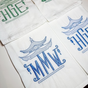 May include: Three white tea towels with blue and green embroidered designs. The towels feature a pagoda design with the letters "MMW" and "HGE" embroidered below.