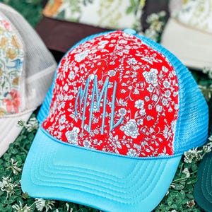 May include: A red and white floral print trucker hat with a light blue brim and mesh back. The hat features the embroidered initials "MML" in light blue. Other hats with floral patterns are visible in the background.