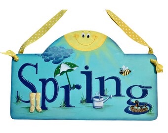 May include: A blue wooden sign with the word "Spring" painted in navy blue. The sign has a yellow sun with a smiling face, a blue cloud with raindrops, a green umbrella, a yellow watering can, a bee, and a bird's nest with a brown bird.