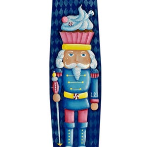 May include: A blue and pink painted nutcracker figurine with a candy cane and cupcake on its head. The nutcracker is holding a candy cane-decorated staff. The background is a blue and white diamond pattern.