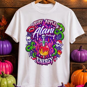 Pode incluir: Uma t-shirt branca com uma estampa gráfica de uma maçã vermelha atravessada por uma adaga, rodeada por chamas roxas, cobras verdes, crânios, teias de aranha e lábios vermelhos. O texto diz Ruby Apple Alani NY Energy em uma fonte estilizada.