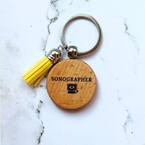 May include: A round wooden keychain featuring the text SONOGRAPHER and a small ultrasound monitor icon. The keychain includes a silver metal ring and a bright yellow faux leather tassel attached to the side of the wooden disc.