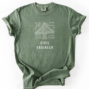 May include: A sage green short-sleeve t-shirt featuring a white line-art graphic of a bridge structure over a grid pattern. Below the graphic, the text CIVIL ENGINEER is printed in white capital letters. The shirt has a crew neckline.