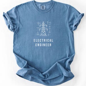 May include: A blue short-sleeved t-shirt featuring a white graphic of an electrical transmission tower centered on the chest. Below the tower, the text reads ELECTRICAL ENGINEER in white capital letters. The shirt has a crew neckline.