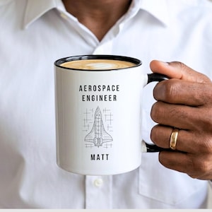 May include: White ceramic mug with a black rim and handle, featuring the text "AEROSPACE ENGINEER" above a line drawing of a space shuttle and the name "MATT". The mug is filled with a light brown liquid.