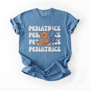 May include: A blue short-sleeve t-shirt featuring the word PEDIATRICS repeated in a wavy white font. Centered on the shirt is a brown teddy bear illustration with a red stethoscope draped around its neck, positioned over the text graphic.