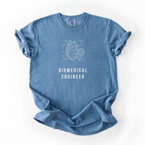 May include: A blue short-sleeve t-shirt featuring a white line-art graphic of an anatomical heart, a gear, and a small heart symbol. Below the graphic, the text BIOMEDICAL ENGINEER is printed in white capital letters on the chest area.