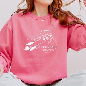 May include: A pink sweatshirt with the text "it's rocket science" and "Aerospace engineer" in white. The design features a rocket ship with a trail of lines. The sweatshirt is a solid pink color.