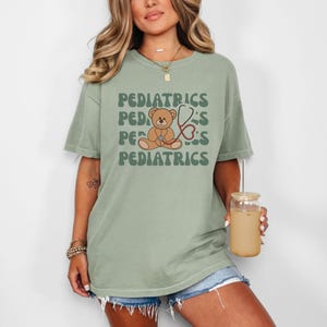 May include: A sage green short-sleeve t-shirt featuring a graphic of a brown teddy bear holding a red stethoscope. The word PEDIATRICS is printed in a wavy, dark green font above and below the bear illustration on the front of the garment.