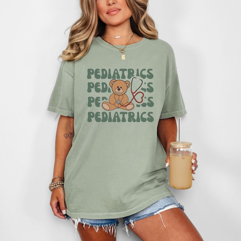 May include: A sage green short-sleeve t-shirt featuring a graphic of a brown teddy bear holding a red stethoscope. The word PEDIATRICS is printed in a wavy, dark green font above and below the bear illustration on the front of the garment.