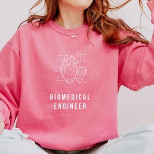 May include: A pink sweatshirt with a white graphic of a heart, gears, and a heart shape. The text "BIOMEDICAL ENGINEER" is printed below the graphic. The sweatshirt has long sleeves and a crew neck.