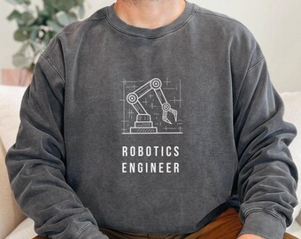 May include: Dark gray sweatshirt featuring a white line drawing of a robotic arm within a grid. Below the graphic, the text reads "ROBOTICS ENGINEER" in a clean, sans-serif font. The sweatshirt has a crew neck and long sleeves, with a slightly faded, vintage look.
