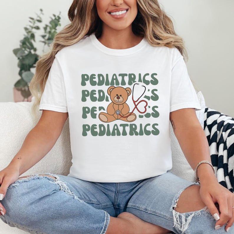 May include: A white t-shirt featuring a brown teddy bear holding a red stethoscope. The word PEDIATRICS is printed in a wavy, sage green font four times in the background, creating a layered graphic design on the front of the garment.