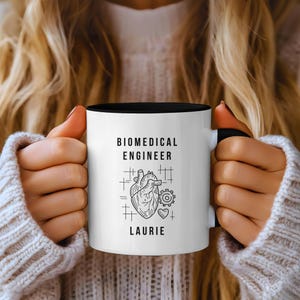 May include: White ceramic mug with a black interior, featuring the text "BIOMEDICAL ENGINEER" above a heart and gear graphic, with the name "LAURIE" below. The mug is held by a person wearing a cream-colored sweater.