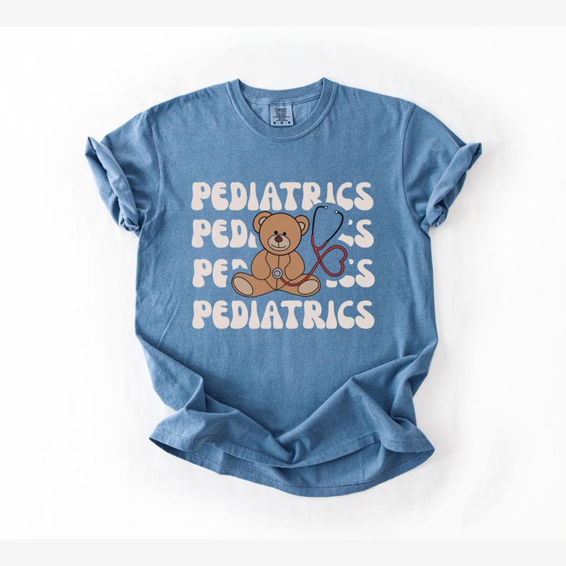 May include: A blue short-sleeve t-shirt featuring the word PEDIATRICS repeated in a wavy white font. Centered on the shirt is a brown teddy bear illustration with a red stethoscope draped around its neck, positioned over the text graphic.
