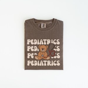 May include: A brown crewneck t-shirt featuring the word PEDIATRICS printed in a wavy, cream-colored font across the chest. A brown teddy bear illustration is centered on the shirt, wearing a red stethoscope that forms a heart shape.