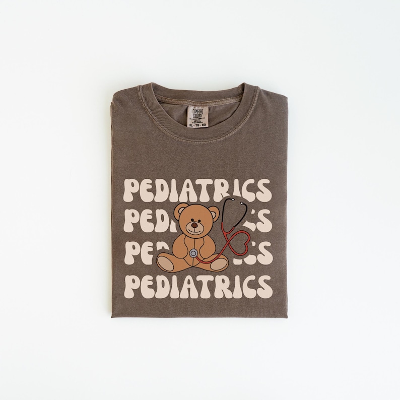 May include: A brown crewneck t-shirt featuring the word PEDIATRICS printed in a wavy, cream-colored font across the chest. A brown teddy bear illustration is centered on the shirt, wearing a red stethoscope that forms a heart shape.