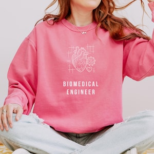 May include: A bright pink long-sleeve sweatshirt featuring a white line-art graphic of an anatomical heart, a small gear, and a heart symbol. Below the graphic, the text reads BIOMEDICAL ENGINEER in a clean, white, sans-serif capital font.