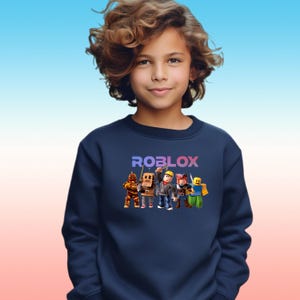 Pu&ograve; includere: Felpa blu navy con la scritta "ROBLOX" in un gradiente viola e blu. Sotto il testo ci sono personaggi dei cartoni animati. La felpa &egrave; indossata da una persona con i capelli castani ricci.
