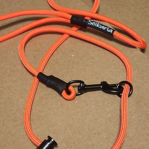 Könnte beinhalten: Leuchtend orangefarbene Hundeleine mit schwarzen Beschlägen. Die Leine besteht aus einem dicken, gewebten Seilmaterial. Der Markenname "Seikaria" ist weiß auf einem schwarzen Band aufgedruckt. Mit Metallverschluss und Handschlaufe.