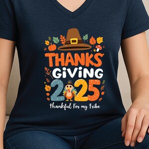 May include: Heather navy v-neck t-shirt with a Thanksgiving graphic. The design includes a pilgrim hat, pumpkins, and the words "THANKS GIVING 2025" with a turkey and the phrase "thankful for my tribe."