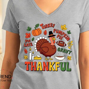 May include: A gray V-neck t-shirt with a colorful Thanksgiving-themed graphic. The graphic includes a turkey, pumpkin, football, and the word "THANKFUL". The shirt is made of Athletic Heather material.