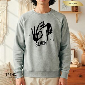 K&ouml;nnte beinhalten: Ein hellgraues Sweatshirt mit einem schwarzen Aufdruck, der Handgesten und die Worte "SIX SEVEN" zeigt. Das Sweatshirt hat einen Rundhalsausschnitt und lange &Auml;rmel. Der Text und die Handgesten sind in einer fetten, serifenlosen Schrift.