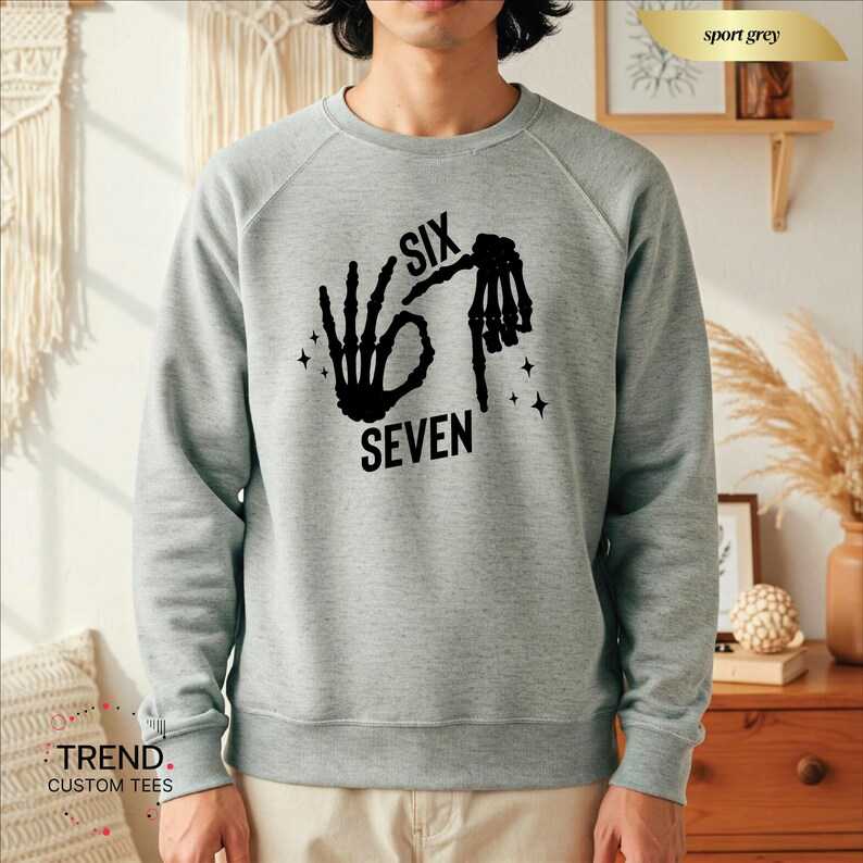 K&ouml;nnte beinhalten: Ein hellgraues Sweatshirt mit einem schwarzen Aufdruck, der Handgesten und die Worte "SIX SEVEN" zeigt. Das Sweatshirt hat einen Rundhalsausschnitt und lange &Auml;rmel. Der Text und die Handgesten sind in einer fetten, serifenlosen Schrift.