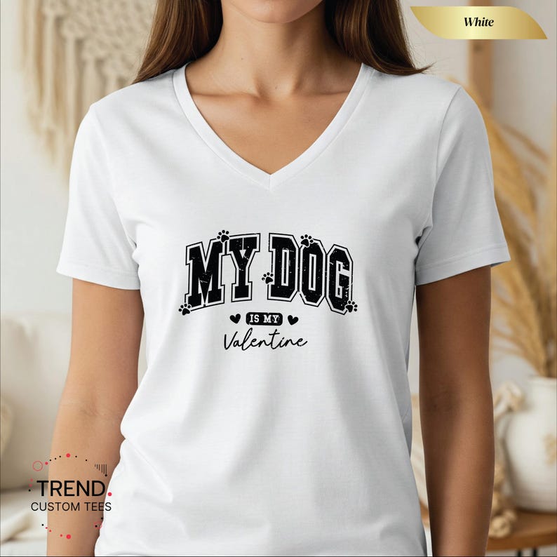 May include: White V-neck t-shirt with the text "MY DOG is my Valentine" in black. The text is in a bold, collegiate-style font, with paw prints and hearts. The shirt is a casual, everyday wear item.