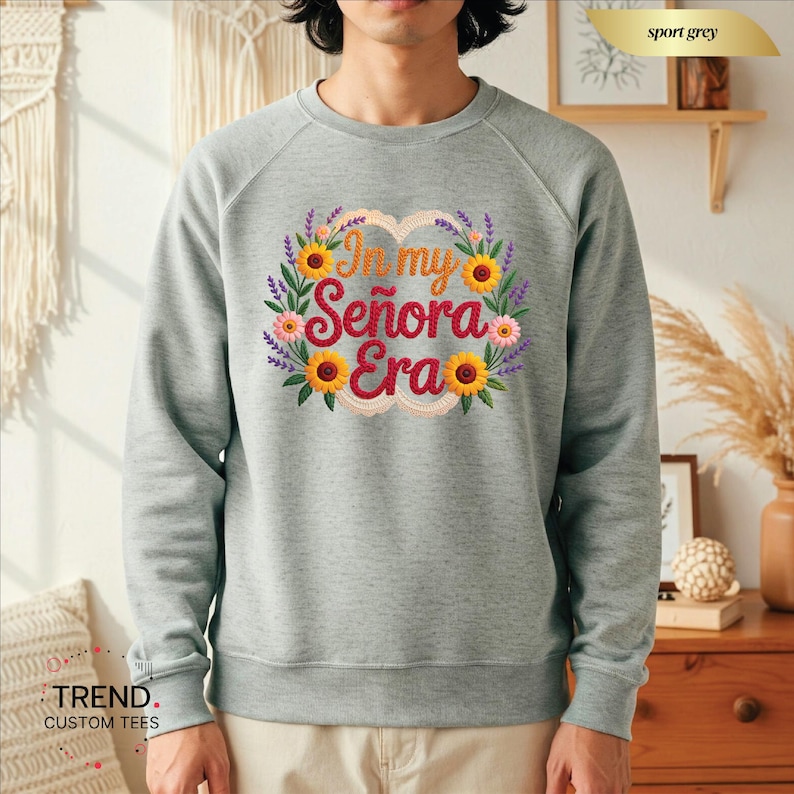 Op de afbeelding: Een lichtgrijs sweatshirt met een bloemenontwerp en de tekst "In my Se&ntilde;ora Era". Het ontwerp bevat zonnebloemen, lavendel en roze bloemen. De sweatshirt heeft een ronde hals.