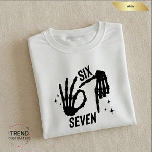 K&ouml;nnte beinhalten: Wei&szlig;es T-Shirt mit einem schwarzen Grafikdesign einer Skeletthand, die das "OK"-Zeichen formt, mit den W&ouml;rtern "SIX" und "SEVEN". Das T-Shirt hat einen Rundhalsausschnitt und ist gefaltet. Das Markenlogo "TREND CUSTOM TEES" ist sichtbar.