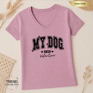 May include: Heather orchid V-neck t-shirt with the text "MY DOG is my Valentine" in black. The text includes paw prints and hearts. The shirt is a light purple color.