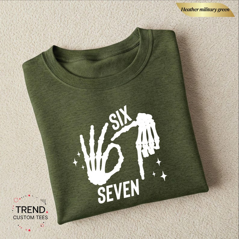 K&ouml;nnte beinhalten: Heather milit&auml;rgr&uuml;nes T-Shirt mit einem wei&szlig;en Aufdruck von Skeletth&auml;nden, die die Zahlen sechs und sieben bilden. Die W&ouml;rter "SIX" und "SEVEN" sind ebenfalls wei&szlig;. Das T-Shirt ist von Trend Custom Tees.