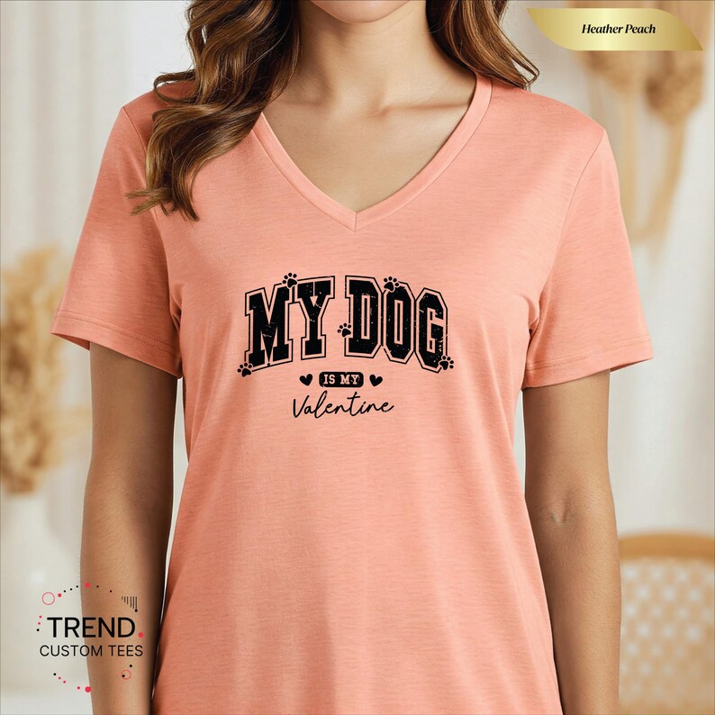 May include: Heather peach V-neck t-shirt with the text "MY DOG is my Valentine" in black. The text is in a collegiate style font with paw prints and hearts. The shirt is a solid color.