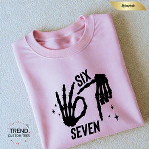 K&ouml;nnte beinhalten: Hellrosa Sweatshirt mit schwarzer Skeletthand-Grafik, die die Zahlen sechs und sieben bildet. Der Text "SIX SEVEN" ist schwarz gedruckt. Das Sweatshirt hat einen Rundhalsausschnitt und lange &Auml;rmel.