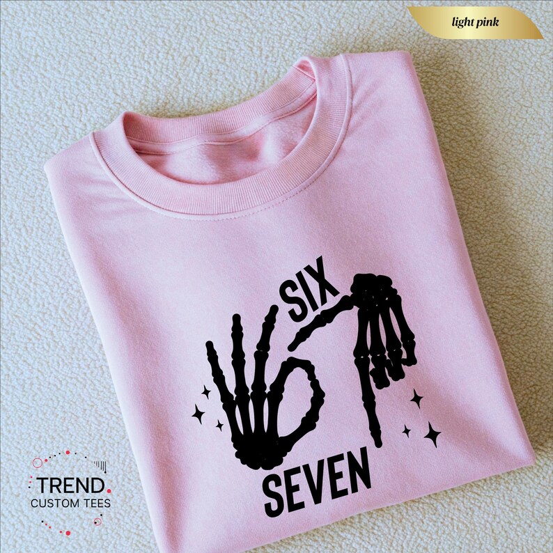 K&ouml;nnte beinhalten: Hellrosa Sweatshirt mit schwarzer Skeletthand-Grafik, die die Zahlen sechs und sieben bildet. Der Text "SIX SEVEN" ist schwarz gedruckt. Das Sweatshirt hat einen Rundhalsausschnitt und lange &Auml;rmel.