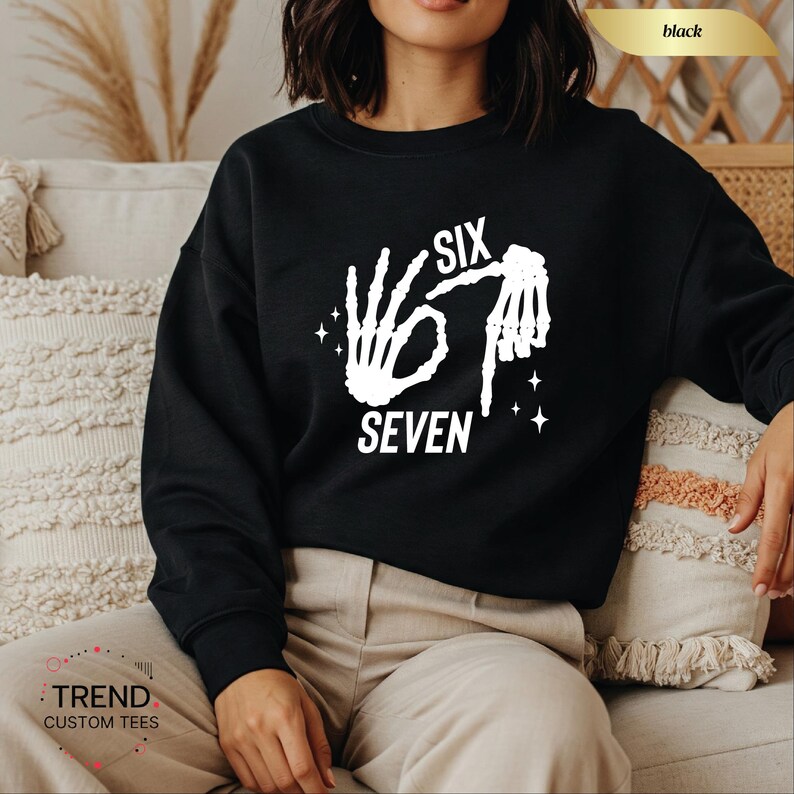 K&ouml;nnte beinhalten: Schwarzes Sweatshirt mit wei&szlig;em Grafikdesign, das die Worte "SIX SEVEN" und eine Skeletthand zeigt, die die Zahl sechs bildet. Das Sweatshirt ist ein Rundhals-Stil und besteht aus einem weichen Material. L&auml;ssiger Stil.